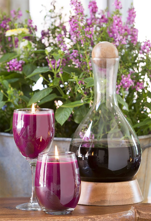 Stemless and stemmed wine glass candles next to a decanter of red wine in front of a planter of purple flowers.