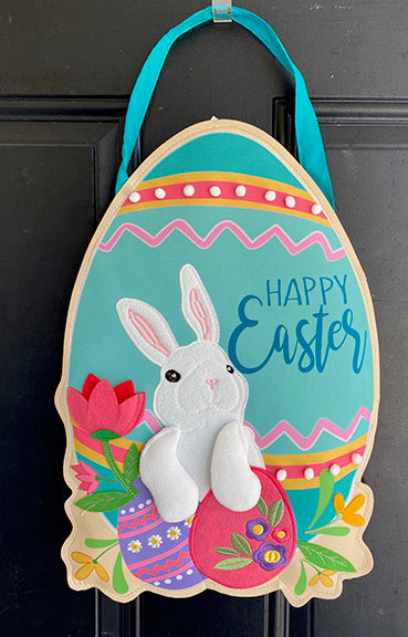 An egg shaped door decoration hanging from a blue ribbon in front of a black door.  The egg itself features dimensional cotton decorations and shows a bunny holding two embroidered eggs.  To the right of the bunny are the words "Happy Easter"