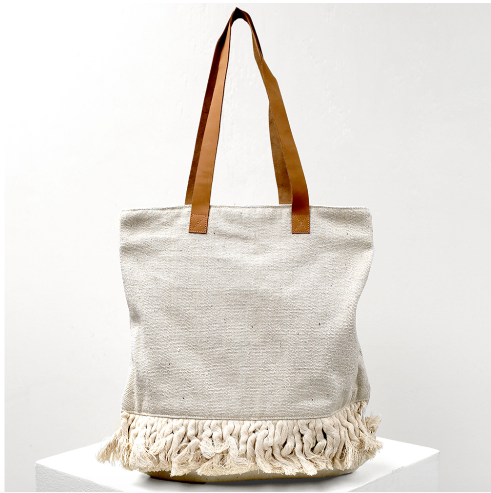 A cotton tote bag on a bag hanger on a white background.  The tote has brown faux leather straps, and the body of the bag is light gray with dark specks throughout.  Along the bottom of the bag is a row of wide braided fringe.