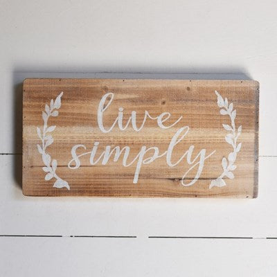 Wooden sign with white text and laurel silhouettes on either side of the text.  The cursive test says in all lower case 'live simply'