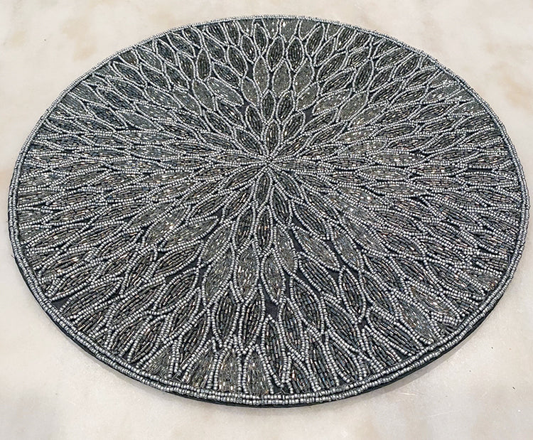 View from above of a glass beaded placemat with a floral pattern.  Beads are in shades of gray and silver.  Round placemat sits on top of a marble surface.