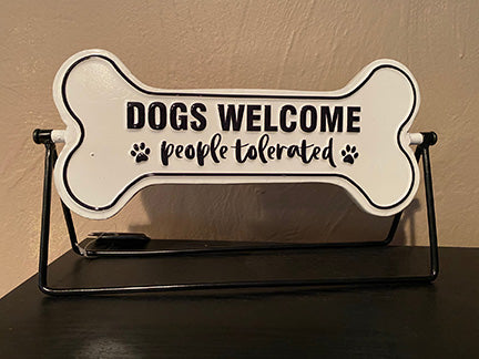 White enamel bone shaped sign on a black metal stand.  Sign can rotate to show a different saying.  Saying on this side is 'Dogs Welcome people tolerated'