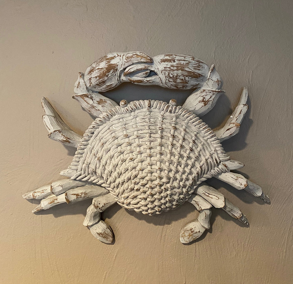 A resin sculpture of a crab hung on a tan wall.  The crab is an off white finish and is weather worn to expose a wood texture below.  The crab shell is carved to look like woven wicker.