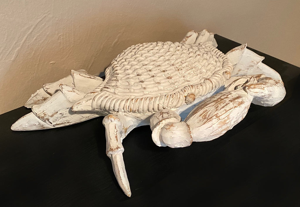A resin sculpture of a crab on a black surface.  The crab is a white washed weathered finish that exposes a wooden finish in parts.  The shell of the crab is carved to look like woven wicker.