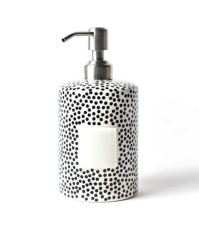 A hand pump soap dispenser on a white background.  The pump itself is polished chrome and the body of the container is a white cylinder with a random black polka dot pattern.  The front of the cylinder has a rectangular square  piece of velcro for decorative attachments.