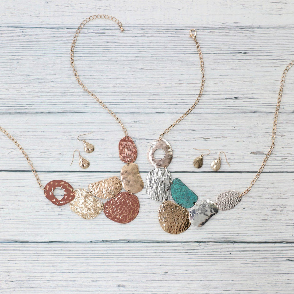 Two necklace and earring sets on a white plank background.  The pendant of each necklace is made of the same shapes but are in different finishes.  The left necklace has tones of gold, and three parts are brushed over with deep red.  The right necklace has mostly silver parts, but 1 is gold and one is turquoise.  Each necklace also features a coordinating pair of earrings shaped like a pear.  