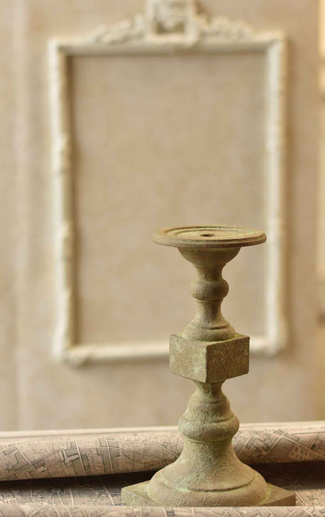 A green candle holder in front of tan architectural features.  The candle holder has indentations for pillars and a hole for a taper.  