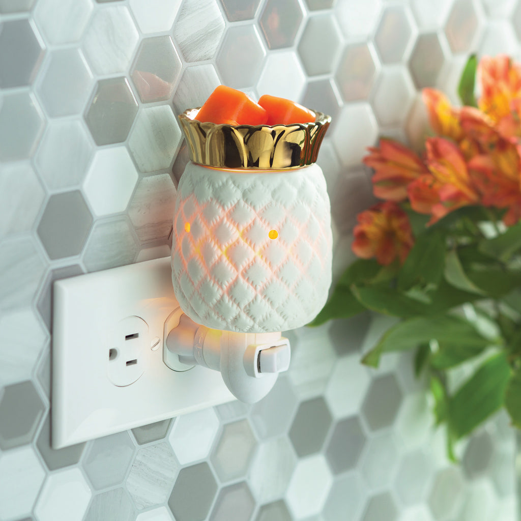 Pineapple wax warmer plugged into an outlet on a wall with hexagon shaped tile.  White ceramic pineapple has a warm internal glow and has gold metallic fronds where the wax sits.