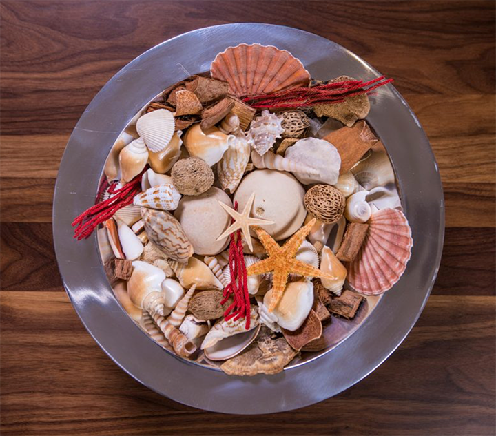 Bowl of potpourri on a wooden table, from above.  Contents of bowl include various shells, starfish, and dried scented items.