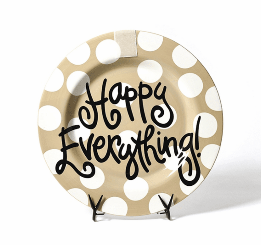 A round platter on a black plate stand in front of a white background.  The plate itself is tan with white polka dots, and has black text in a fun font over the entire platter that says 'Happy Everything!'.  At the top center of the platter is a rectangle of velcro to receive attachments.