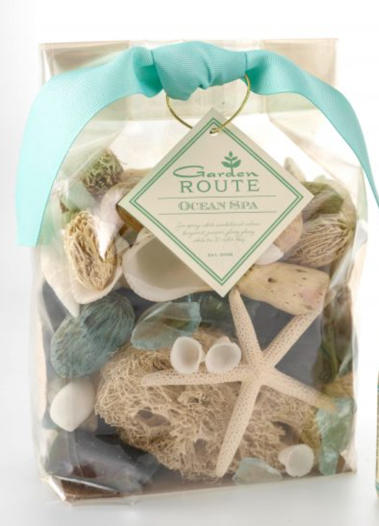 A bag of potpourri on a white background.  Inside the clear bag is an assortment of sponges, dried nuts, shells, and seaglass.  The bag is fastened closed with a light blue ribbon and a tag that says 'Garden Route Ocean Spa'