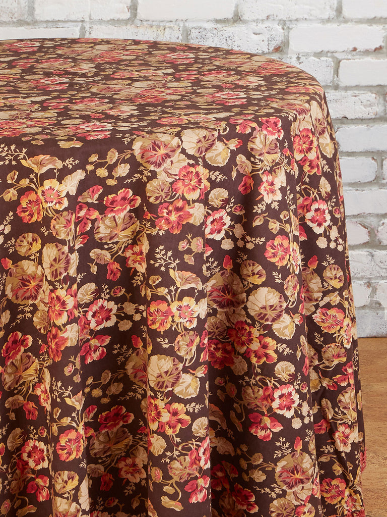 Close up of a floral tablecloth on a round table in front of a white brick wall.  Table cloth has a dark brown background, tan and red flowers, and a woven dark pink bottom detail.