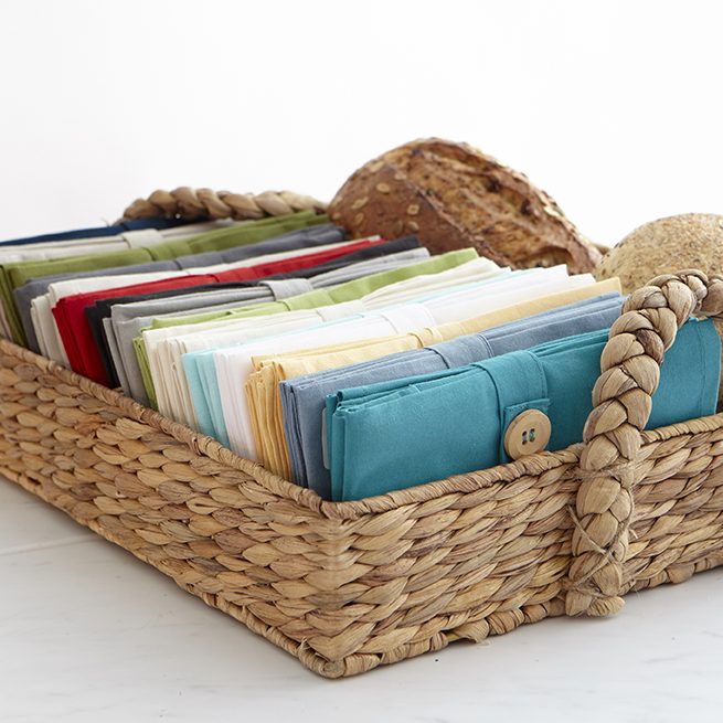 Wicker tray with handles and an assortment of sets of napkins inside.  Each is bundled with a tie featuring a large wooden button.
