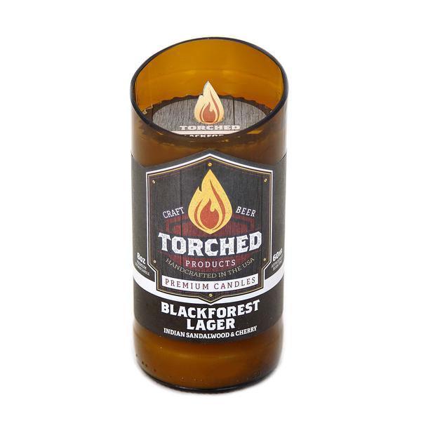 An 8 oz. beer bottle with the top cut off at a 20 degree angle.  The bottle has been filled with wax and is being sold as a candle.  The label shows an illustration of a flame and says 'Torched, craft beer premium candles.  Blackforest Lager, Indian Sandlewood & Cherry'