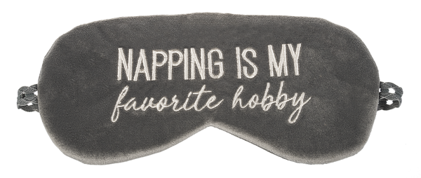 A velvet sleeping mask on a white background.   The mask is gray with off white embroidered text that says 'Napping is my favorite hobby'