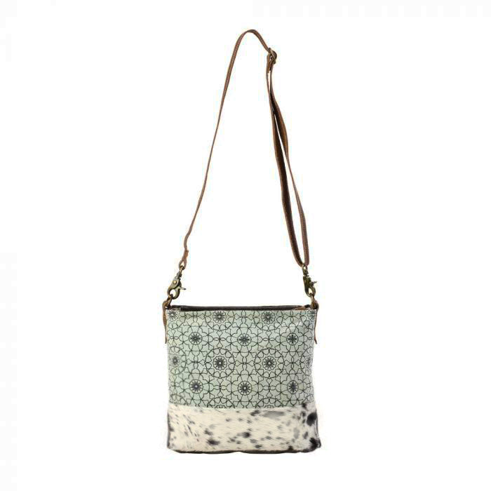 Green and dark gray ogee patterned canvas bag with white and black spotted cow hair trim at the bottom.  A thin dark brown strap is stretched upwards to a hook.