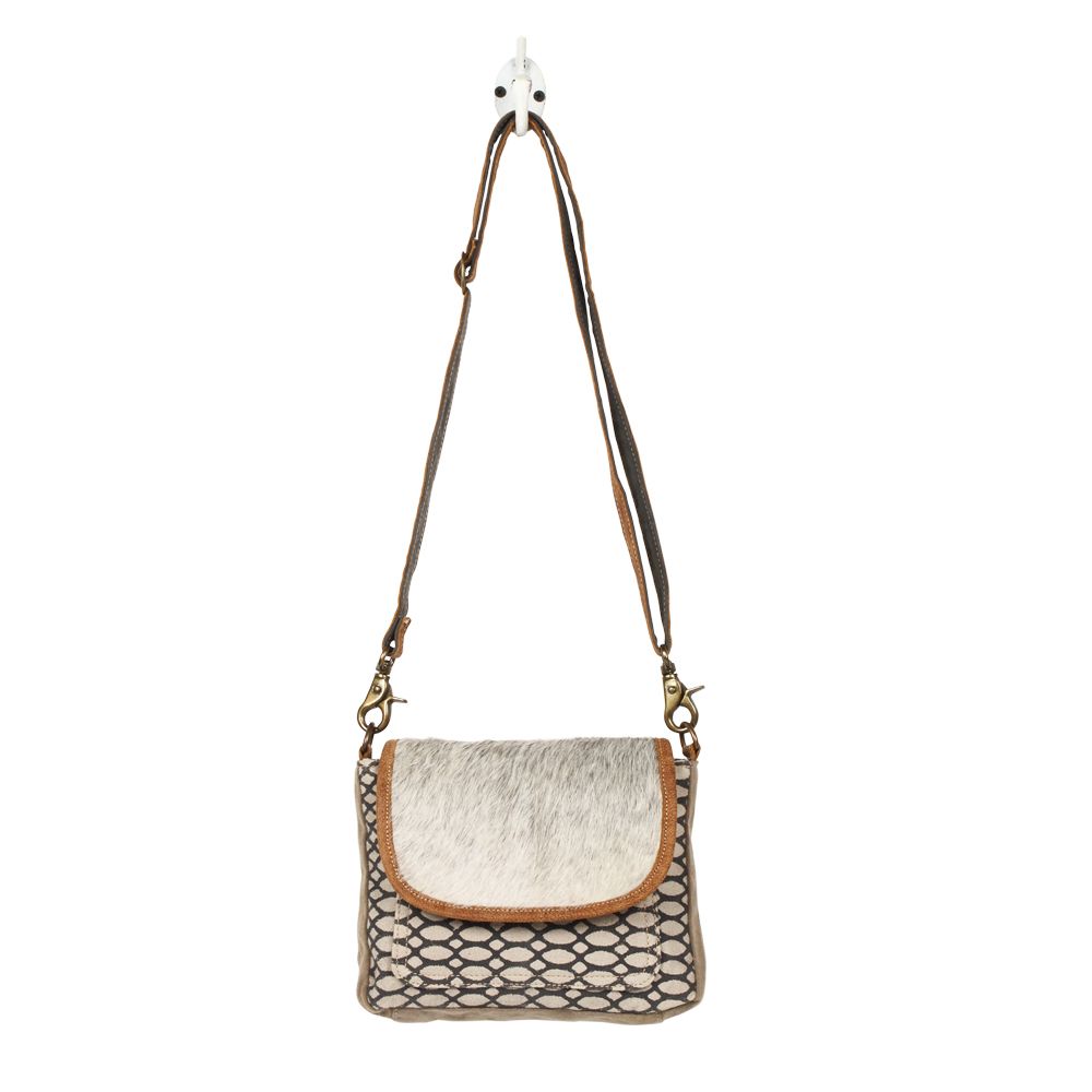 A crossbody bag hung from a wall hook.  The bag has a cowhide flap with a brown leather trim, and the body of the bag is a honeycomb printed pattern on canvas.