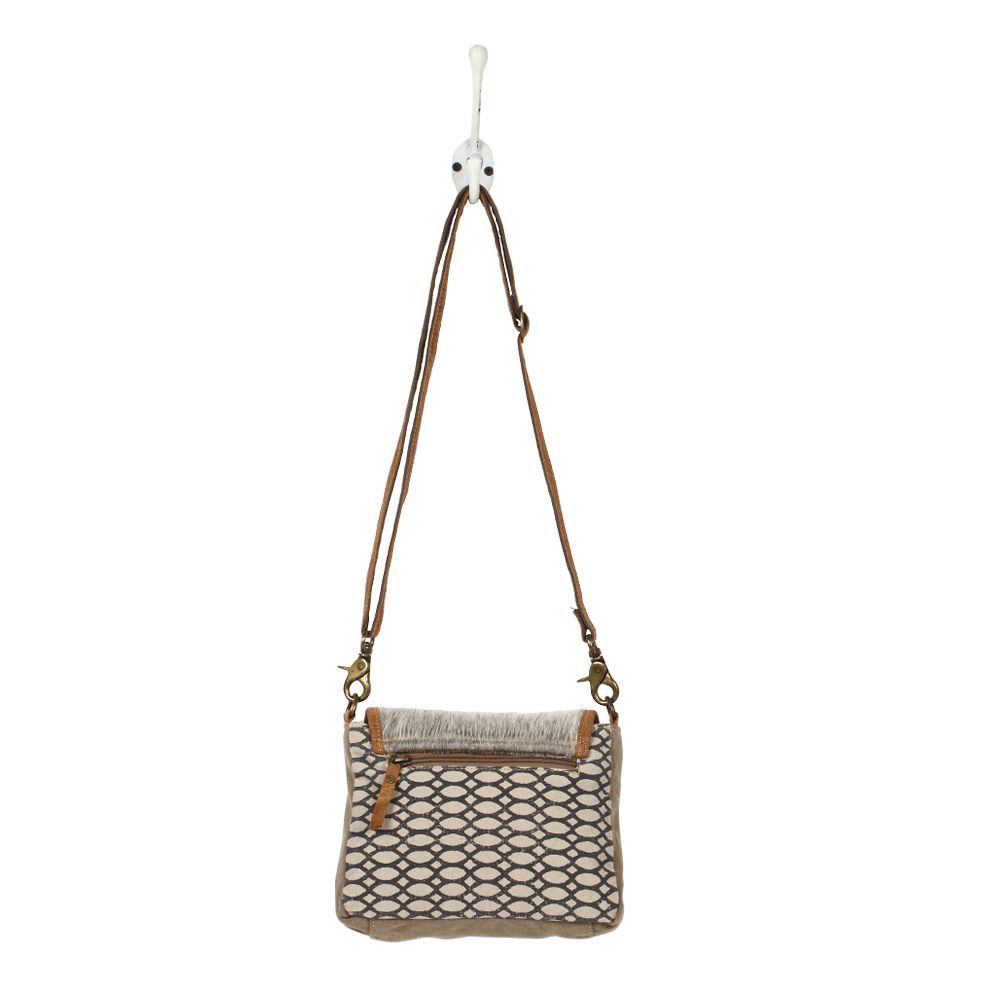 The back view of a crossbody bag hung from a wall hook.  The bag has a cowhide flap with a brown leather trim, and the body of the bag is a honeycomb printed pattern on canvas.  There is a zippered compartment towards the top of the bag.