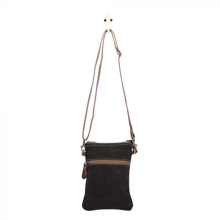 A photo of the back of a crossbody bag.  It is a mottled black, and has a horizontal brown zipper 1/4 of the way down from the top.  The strap is stretched upwards towards a white hook.